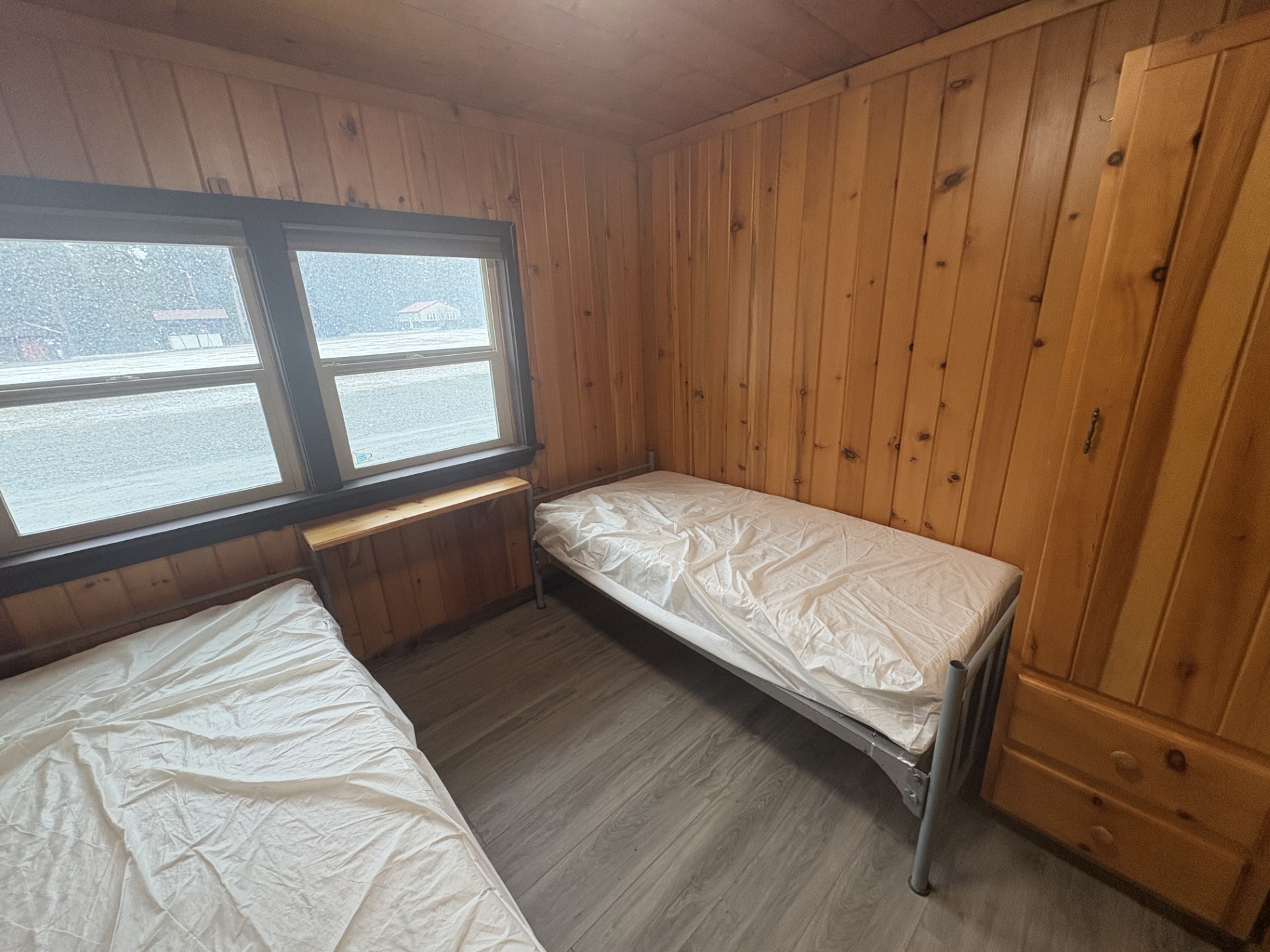 Small wood‑paneled bedroom with two simple twin beds covered in white sheets, one along the right wall and one partially visible at the bottom left, a narrow wooden shelf under a double window, and gray vinyl flooring.