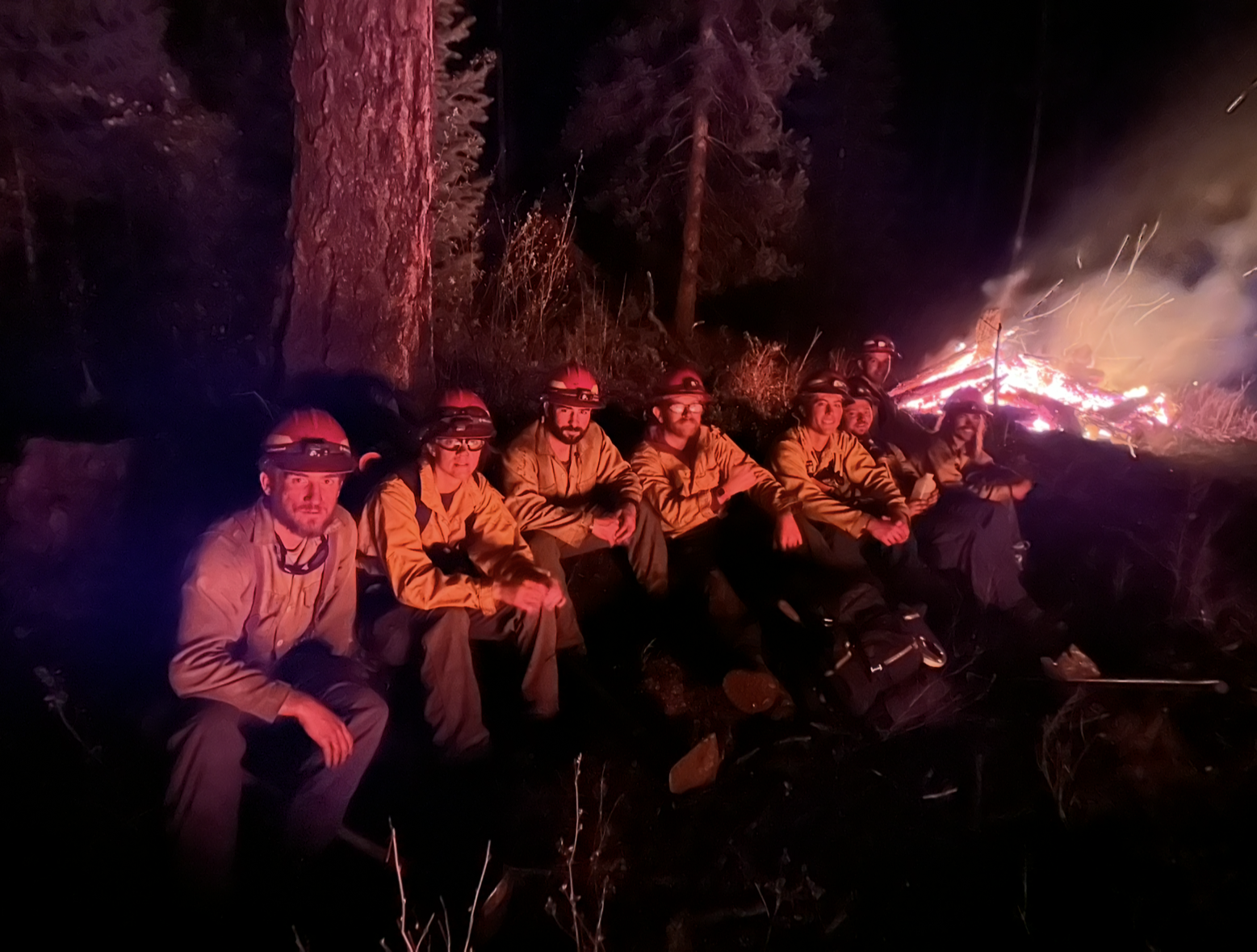 Group of wildland firefighters in yellow shirts and helmets sitting on the ground at night in front of a large glowing burn pile, surrounded by dark trees and illuminated by orange firelight.