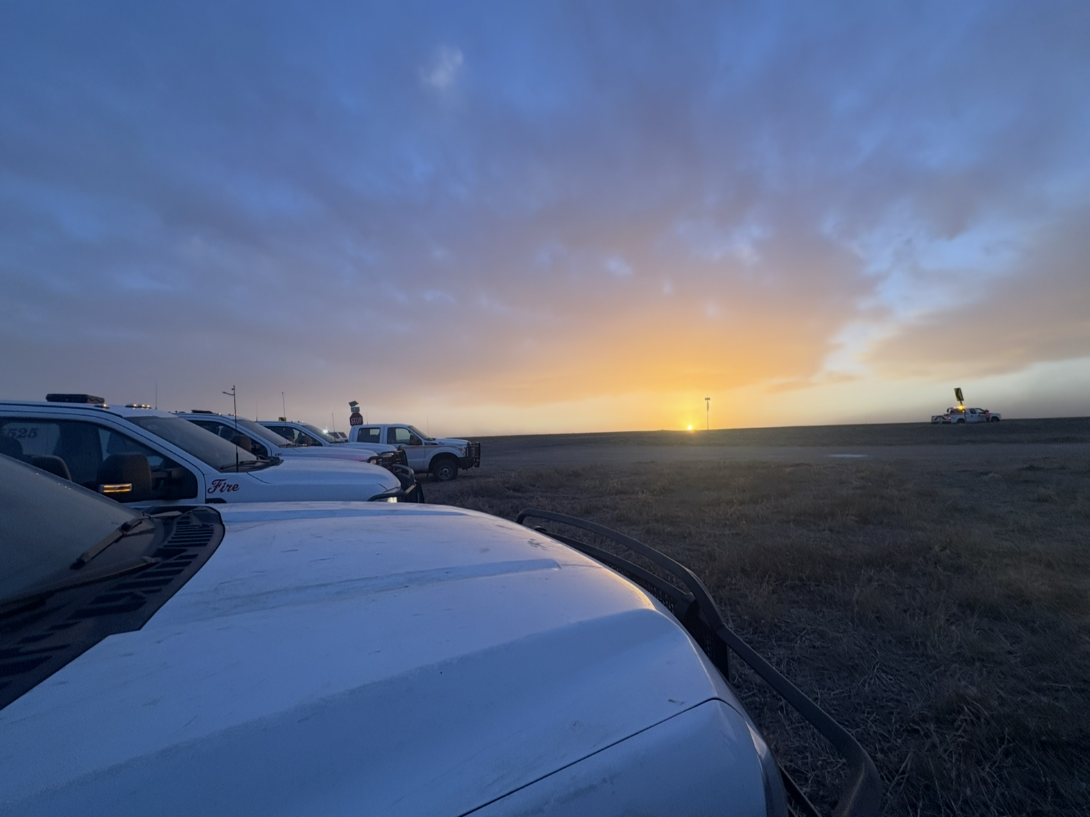 Line of white fire department pickup trucks parked on open prairie at dusk, with a colorful sunset on the horizon and a mostly clear sky above.