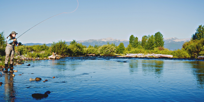 woman fly fishes in river with mountains in the background