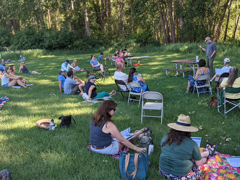 A public meeting outdoors, participants are sitting in the grass listening to the speaker